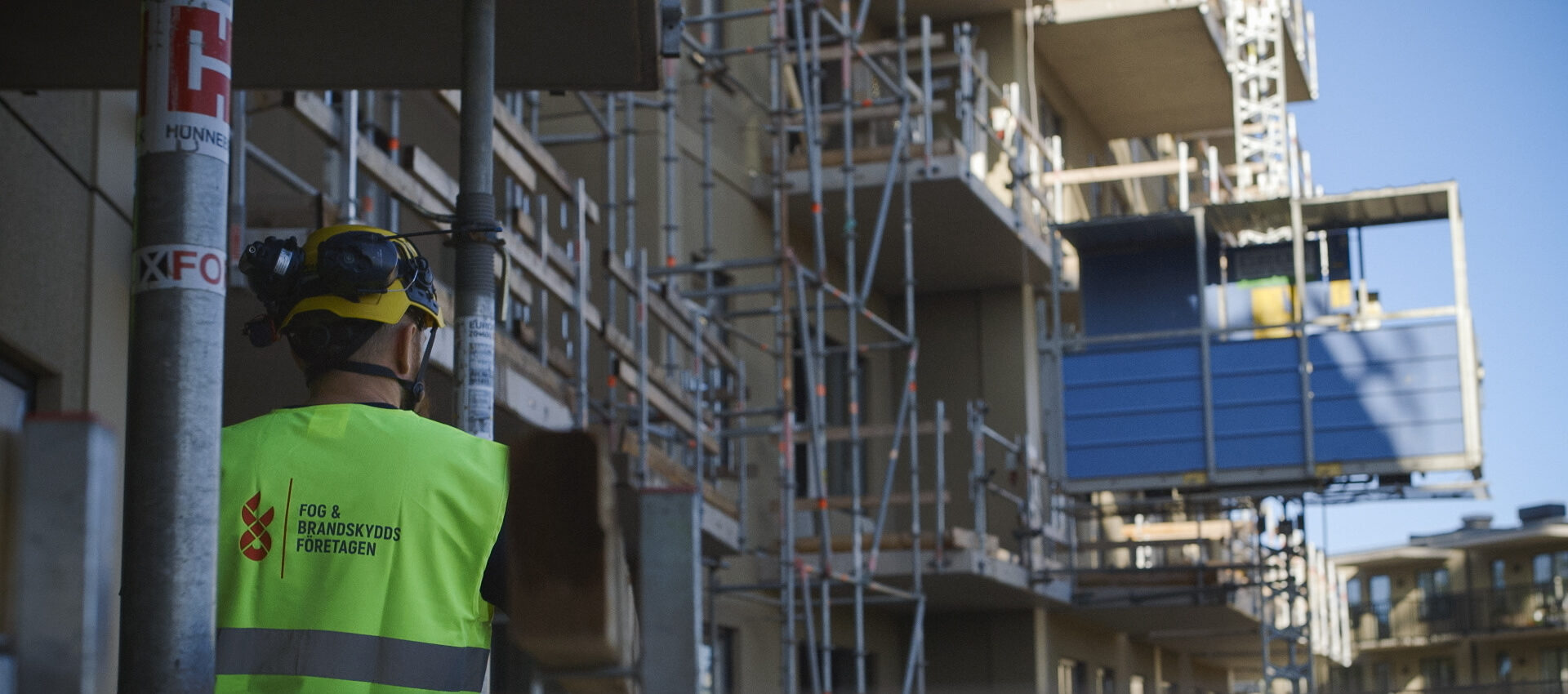 Byggarbetare i hjälm och varselväst från Fog & Brandskydds Företagen står vid en byggarbetsplats med ställningar och pågående nyproduktion i bakgrunden.
