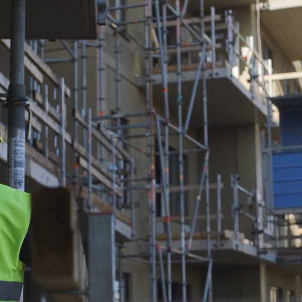 Byggarbetare i hjälm och varselväst från Fog & Brandskydds Företagen står vid en byggarbetsplats med ställningar och pågående nyproduktion i bakgrunden.